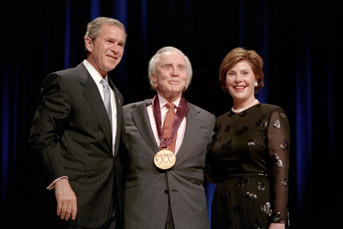 A man in a suit stands with two people, one wearing a black dress, as he receives an award on stage.