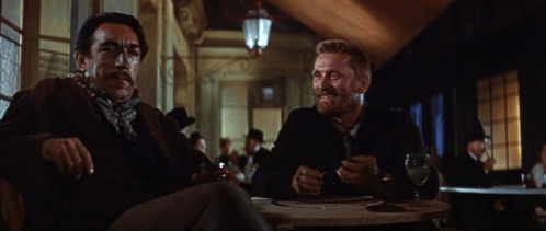 Two men sit at a table in a dimly lit room, engaged in conversation, with drinks in front of them.