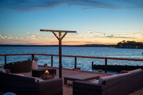 Cozy outdoor seating area by the water at sunset, with string lights and a fire pit.