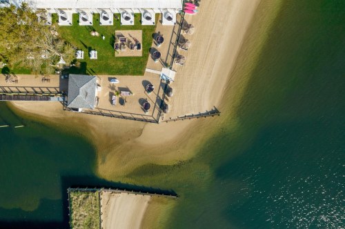 Aerial view of a beachside area with a deck, seating, and water, showcasing sandy shores and green spaces.