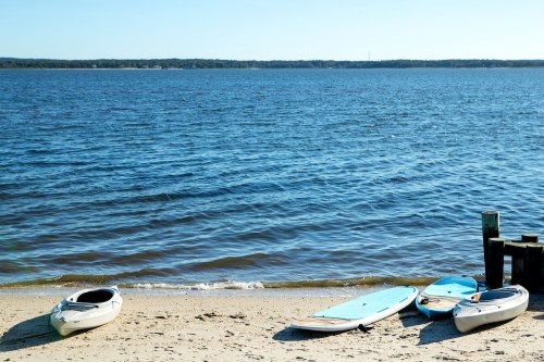 Four kayaks and paddleboards on a sandy beach by a calm blue lake under a clear sky.
