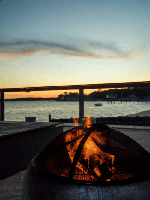 A cozy fire pit glows against a serene sunset over the water, with boats in the distance and a calm sky.