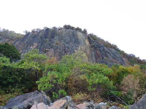 A rocky cliff rises above a forest of green and autumn-colored trees under a cloudy sky.