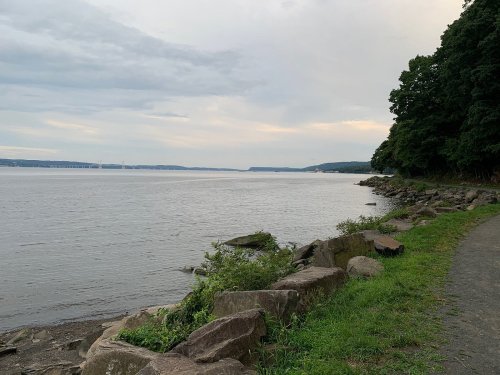 A serene riverside view with rocky shorelines, lush greenery, and a cloudy sky in the background.