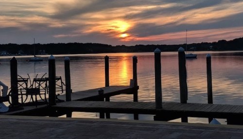 Sunset over a calm lake, reflecting orange and purple hues, with a wooden dock and moored boats in the background.