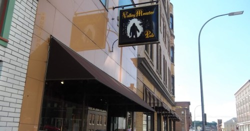 Sign for Valley Monster Pub hanging outside a building, with a reflection of the street visible on the glass.
