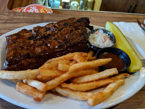 A plate with BBQ ribs, French fries, coleslaw, a pickle, and two dipping sauces on a wooden table.
