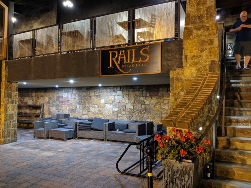 Interior of Rails Steakhouse featuring stone walls, seating area, and a prominent sign. A person is walking up the stairs.