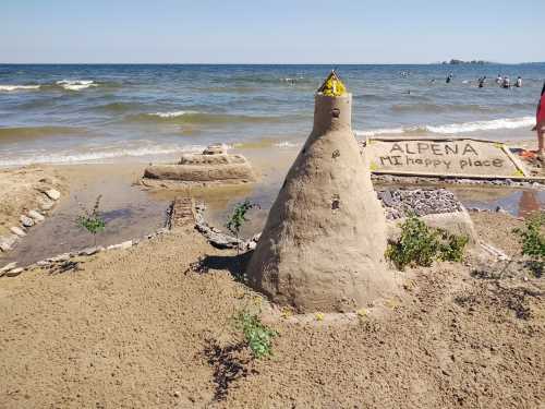 A sandy beach scene featuring a large sandcastle with a flag, surrounded by smaller structures and a lake in the background.