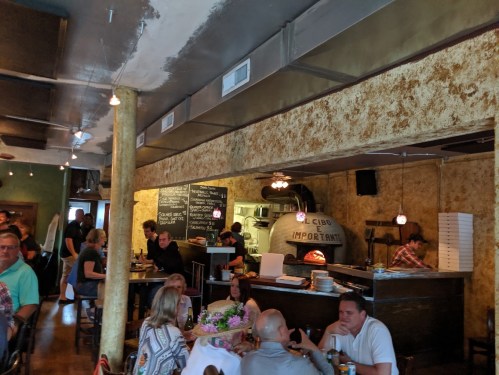 A bustling restaurant interior with diners, a bar area, and a visible kitchen in the background. Warm, inviting atmosphere.