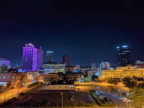 Night view of a city skyline with illuminated buildings and a dark sky, featuring vibrant purple lights.