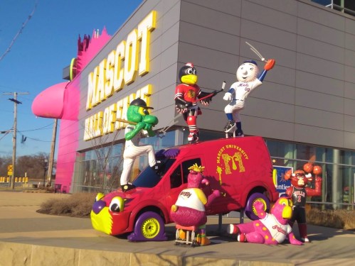 Colorful mascots in various sports poses atop a van outside the Mascot Hall of Fame building.