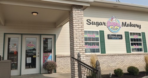 Exterior of Sugar Sugar Makery, featuring a sign and windows with colorful displays of candy and sweets.