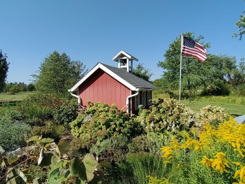 A red building with a bell tower surrounded by lush greenery and flowers, with an American flag waving nearby.