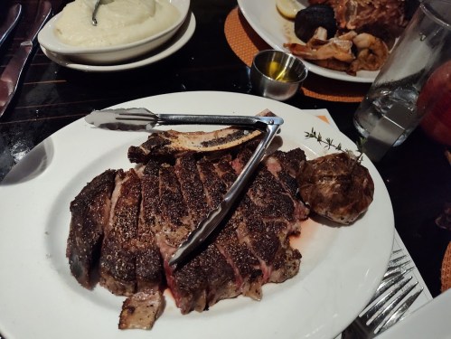 A plate of sliced steak with a baked potato, served with tongs and a small bowl of sauce on the side.