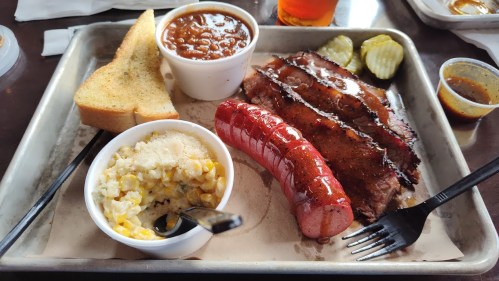 A tray with sliced brisket, sausage, baked beans, corn, pickles, and toast, accompanied by a drink.
