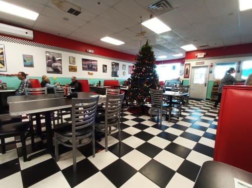 A retro diner with checkered floors, festive decorations, and patrons seated at tables near a Christmas tree.