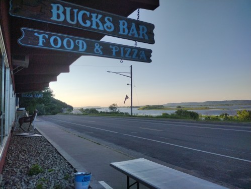 Sign for "Bucks Bar Food & Pizza" with a view of a river and hills at sunset, along a quiet road.