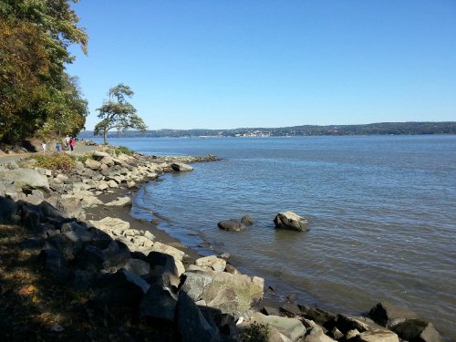 A serene riverside scene with rocky shorelines, trees, and a clear blue sky reflecting on the water.