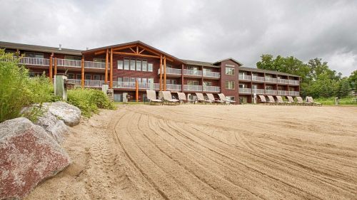 A sandy area in front of a multi-story lodge with lounge chairs and greenery, under a cloudy sky.