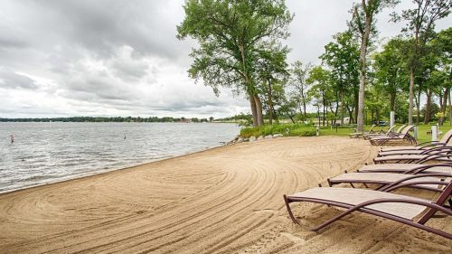 A sandy beach with lounge chairs facing a calm lake, surrounded by trees under a cloudy sky.