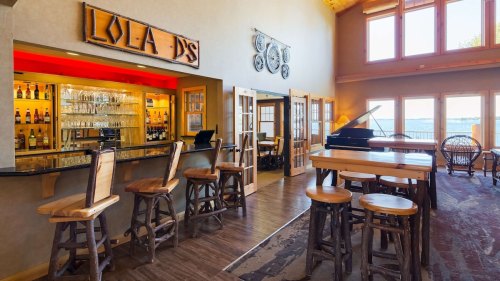 Interior of a bar with wooden stools, a counter, and large windows overlooking a scenic view. Cozy and inviting atmosphere.