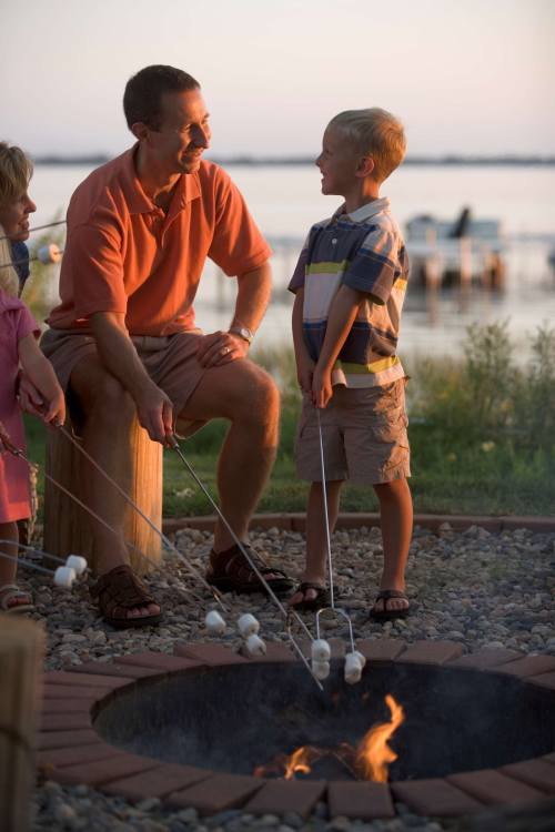 A man and two children roast marshmallows over a fire pit by a lake at sunset.