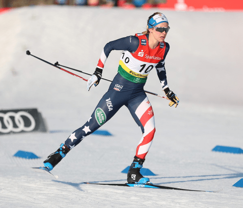 A female cross-country skier in a patriotic outfit races on snow, wearing bib number 10 during a competition.