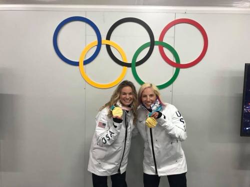 Two smiling athletes in Olympic jackets hold their medals in front of the Olympic rings backdrop.