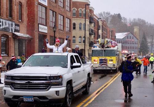 A festive parade scene with people waving from a white truck, snow falling, and a fire truck in the background.
