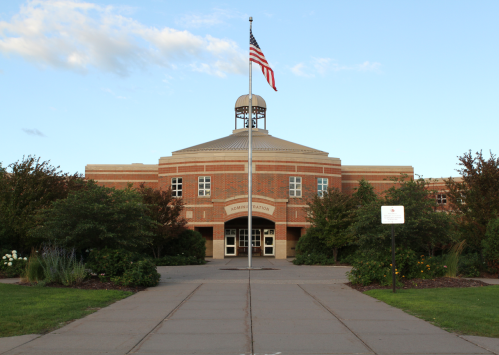 A brick building with a dome and flagpole, surrounded by greenery and a pathway leading to the entrance.