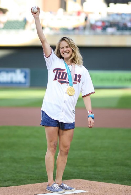 A smiling woman in a Twins jersey raises her arm, holding a gold medal, standing on a baseball field.