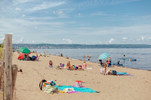 A sunny beach scene with people relaxing on towels, umbrellas, and swimming in calm waters under a blue sky.