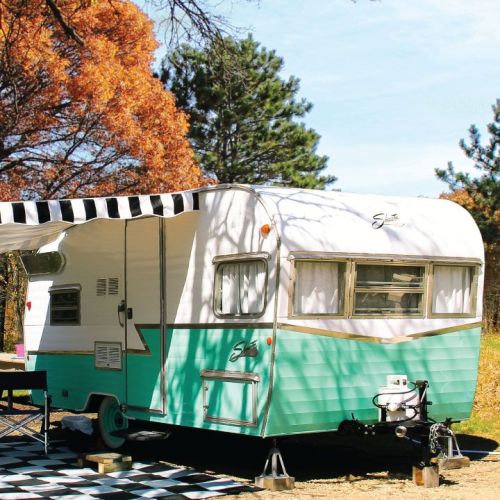 A vintage teal and white camper trailer parked under trees with an awning, set on a black and white checkered ground.