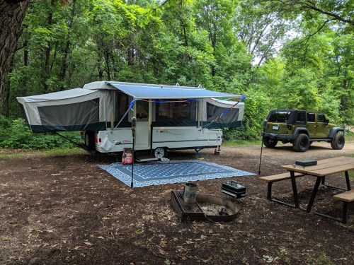 A pop-up camper with an awning is parked in a wooded campsite, next to a Jeep and a picnic table.