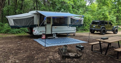 A pop-up camper with an awning set up in a wooded area, next to a Jeep and a picnic table with a fire pit nearby.