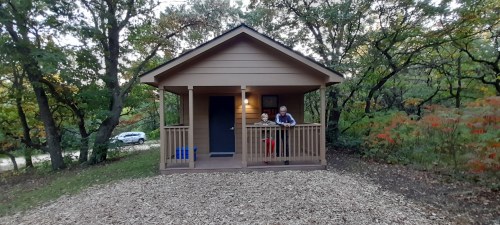 A small cabin surrounded by trees, with two people sitting on the porch and a car parked nearby.