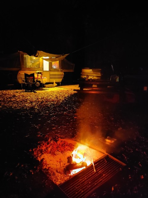 A cozy campsite at night with a glowing fire pit and a camper illuminated by warm light.