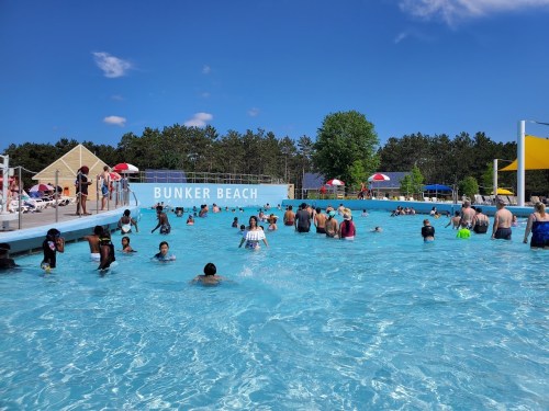 A crowded water park pool on a sunny day, with people swimming and enjoying the water under colorful umbrellas.