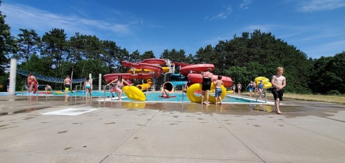 A sunny pool scene with children playing, water slides in the background, and colorful inflatable tubes scattered around.