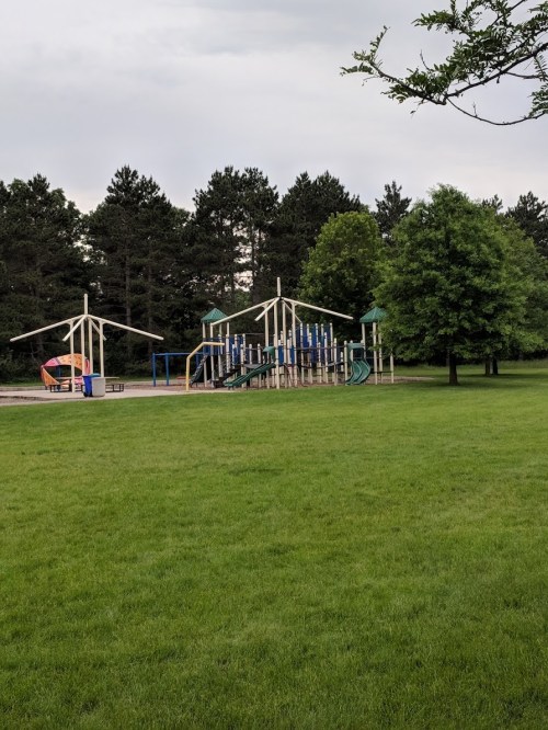 A playground with slides and climbing structures, surrounded by green grass and trees under a cloudy sky.