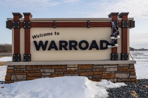 Welcome sign for Warroad, featuring a pair of ice skates hanging on the side, with snow-covered ground in the background.