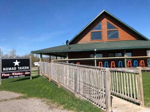 A wooden tavern with a green roof and large windows, featuring a sign that reads "Nomad Tavern" and colorful outdoor seating.