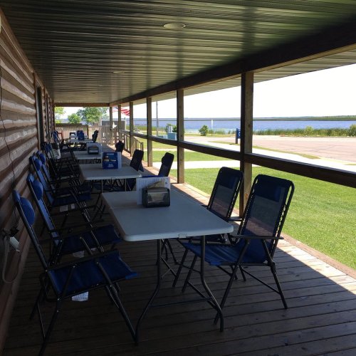 A covered porch with tables and chairs overlooking a calm body of water and green grass.
