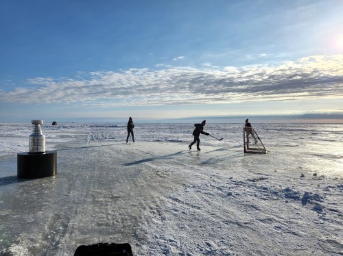 Two people playing hockey on a frozen lake, with the Stanley Cup nearby and a clear blue sky above.