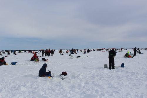 A snowy landscape filled with people ice fishing, some sitting and others standing, under a cloudy sky.