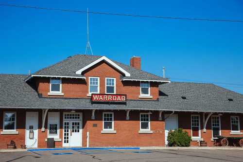 Historic brick train station with a sign reading "Warroad" against a clear blue sky.