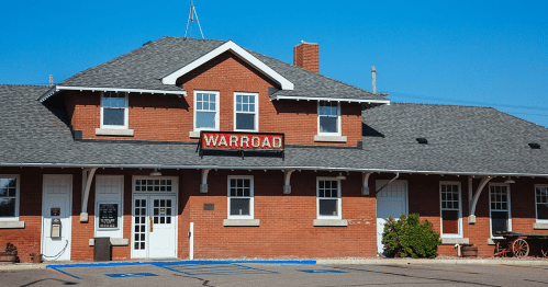 Historic brick building with a gray roof, featuring a sign that reads "WARROAD" and a clear blue sky in the background.
