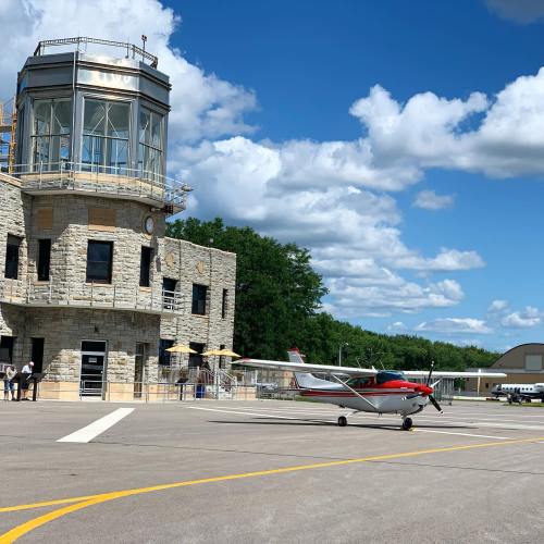 A small red and white airplane is parked near a stone airport control tower under a blue sky with fluffy clouds.