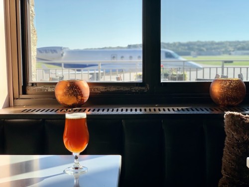 A glass of beer sits on a table by a window, with a private jet visible outside on the runway.